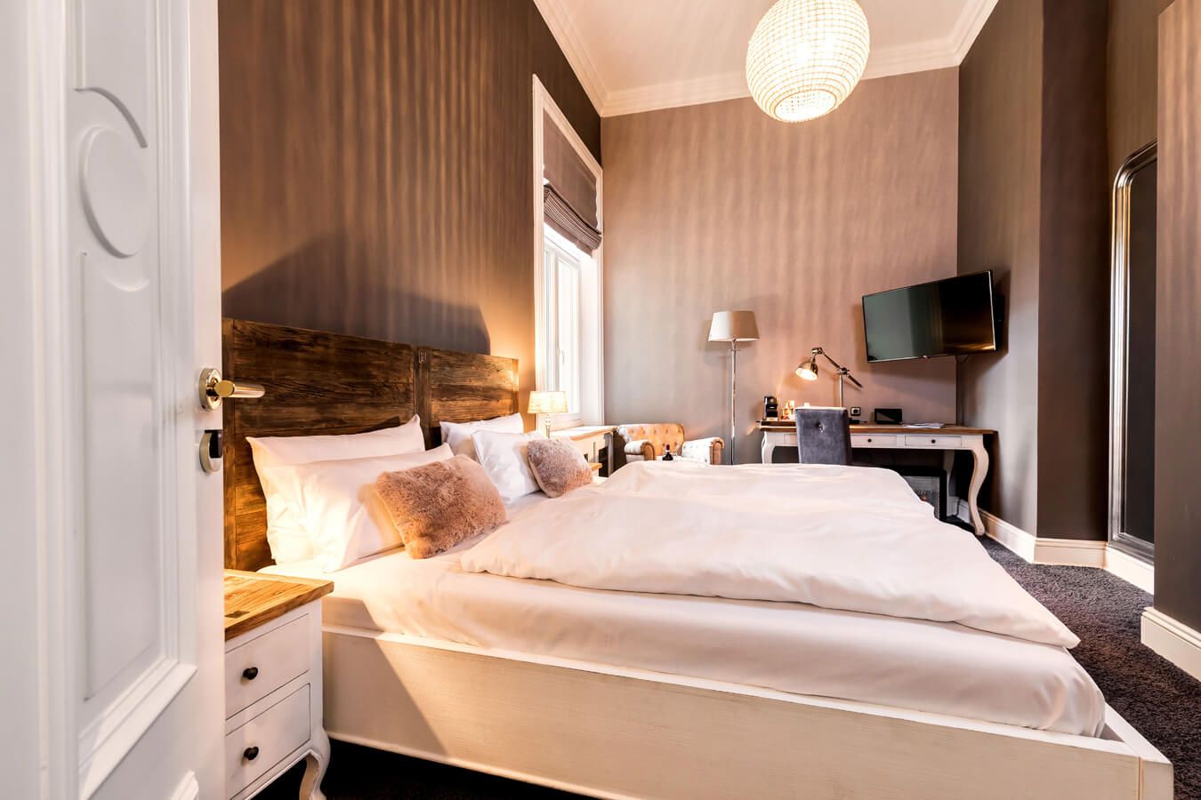 Ein gemütliches Schlafzimmer mit einem großen Bett, weißem Bettzeug, einem Kopfteil aus Holz, Nachttischen, einem Schreibtisch mit Lampe, einem montierten Fernseher, einem Sessel und einem runden Kronleuchter, alles in warmen, neutralen Tönen.