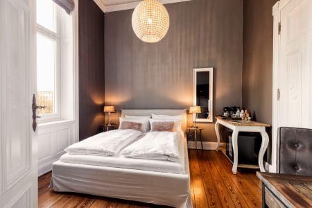 Ein modernes, gemütliches Schlafzimmer mit einem Doppelbett, Holzfußboden, zwei Nachttischlampen, einem großen Fenster mit weißen Vorhängen, einem Kronleuchter, einem Spiegel und einem kleinen Schreibtisch mit einer Kaffeemaschine.