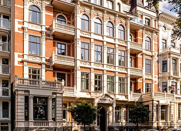 Ein historisches, verziertes Wohnhaus mit großen Fenstern, dekorativen Verzierungen und einer Fassade aus rotem Backstein und cremefarbenem Stein, umgeben von Bäumen und in Sonnenlicht getaucht.