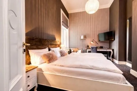 Modernes Schlafzimmer mit großem Bett, flauschiger weißer Bettwäsche, Nachttischen, an der Wand montiertem Fernseher, Schreibtisch mit Stuhl, Lampe und neutral getönten Wänden, beleuchtet von einer runden Deckenleuchte.