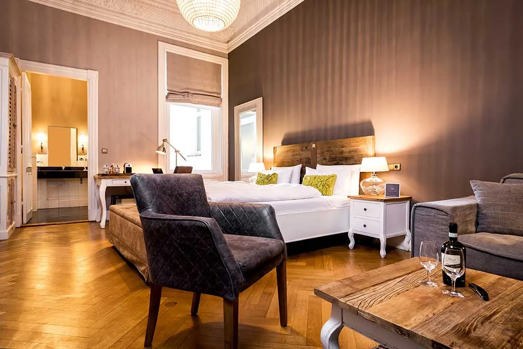 Ein stilvolles Hotelzimmer mit einem großen Bett, zwei Sesseln, einem Sofa, einem hölzernen Couchtisch mit einer Weinflasche und Gläsern, einem Schreibtisch mit einer Lampe und warmer Beleuchtung, mit Holzböden und elegantem Dekor.