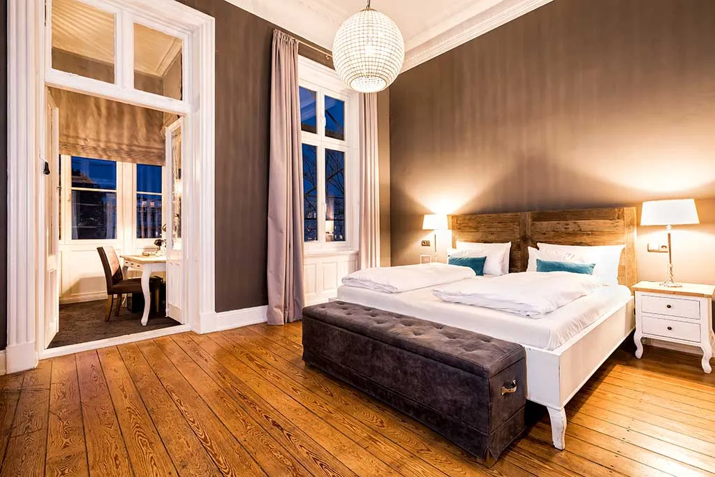 Ein modernes Schlafzimmer mit Holzboden, großem Bett mit weißer Bettwäsche, zwei Nachttischen mit Lampen, einer grau gepolsterten Bank, hohen Fenstern mit Vorhängen und einem Nebenraum, der durch eine offene Doppeltür zu sehen ist.