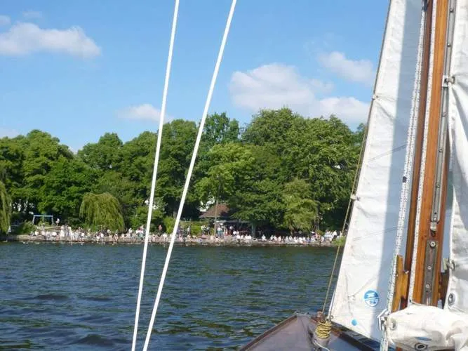 Ein Segelboot auf einem See, dessen Mast und Segel im Vordergrund zu sehen sind, und eine große Menschenmenge, die sich entlang eines von Bäumen gesäumten Ufers unter einem blauen Himmel mit vereinzelten Wolken versammelt hat.