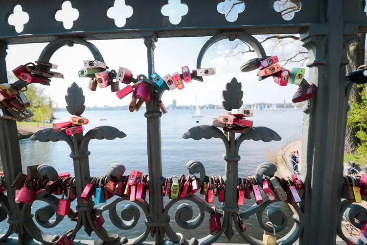Ein mit bunten Vorhängeschlössern versehenes Metallgeländer mit Blick auf einen malerischen Fluss mit Booten und Bäumen im Hintergrund an einem sonnigen Tag.