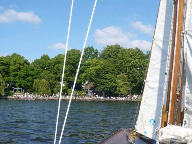 Ein Segelboot auf einem See, dessen Mast und Segel im Vordergrund zu sehen sind, und eine große Menschenmenge, die sich entlang eines von Bäumen gesäumten Ufers unter einem blauen Himmel mit vereinzelten Wolken versammelt hat.