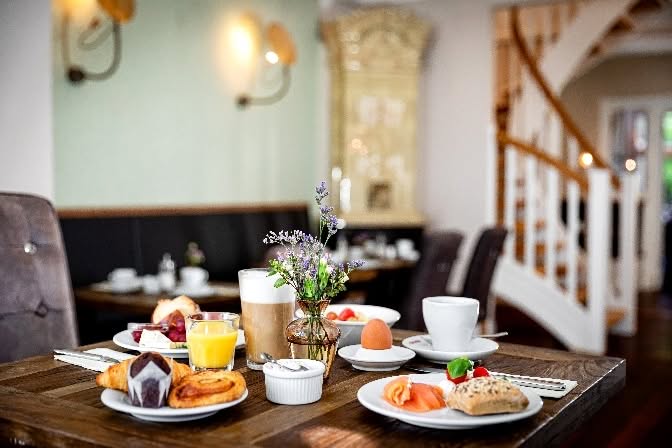 Ein für das Frühstück gedeckter Holztisch mit Kaffee, Saft, Gebäck, Obst, Lachs und einer kleinen Blumenvase. Der Hintergrund zeigt einen gemütlichen, stilvollen Essbereich mit einer Treppe und sanfter Beleuchtung.