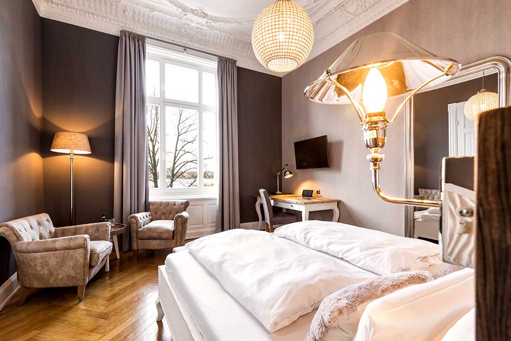 Ein helles, elegantes Schlafzimmer mit weißer Bettwäsche, einem großen Fenster mit grauen Vorhängen, Samtsesseln, einem Schreibtisch mit Fernseher und dekorativer Beleuchtung, mit verschnörkelten Deckenleisten und Holzfußboden.