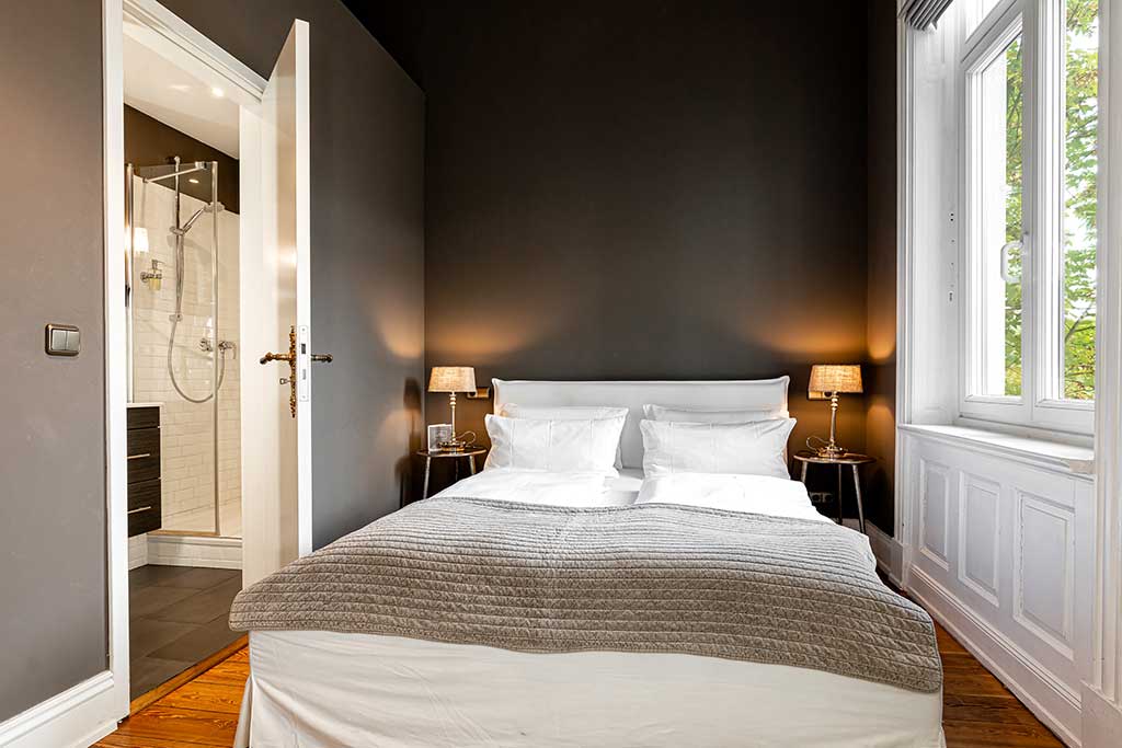 Ein modernes Schlafzimmer mit dunklen Wänden, einem Doppelbett mit weißem Bettzeug und einer grauen Decke, zwei Nachttischen mit Lampen, einem Holzfußboden, einer offenen Tür, die zum Badezimmer führt, und einem großen Fenster, das Tageslicht hereinlässt.